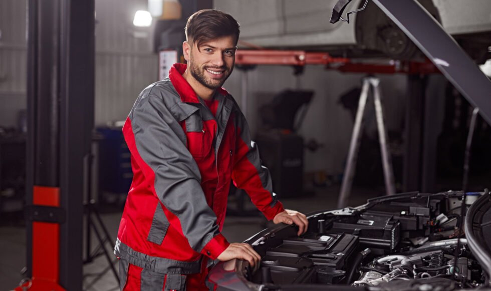 Technician examining car engine in workshop Side view of professional repairman in workwear looking at camera and smiling friendly while standing near car with open hood during diagnostic service in workshop