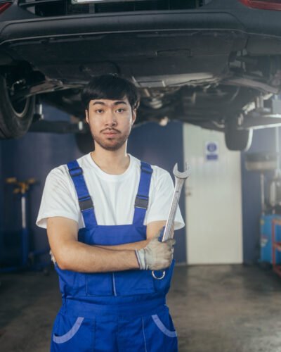 Asian car mechanic worker or repairman holding wrench in arms crossed at auto repair shop looking at camera. After service and transportation career