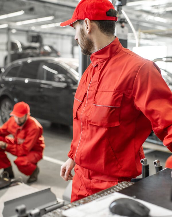 Auto mechanics serving luxury car at the car service Two handsome auto mechanics in red uniform making wheel alignment with professional tools and computer at the car service