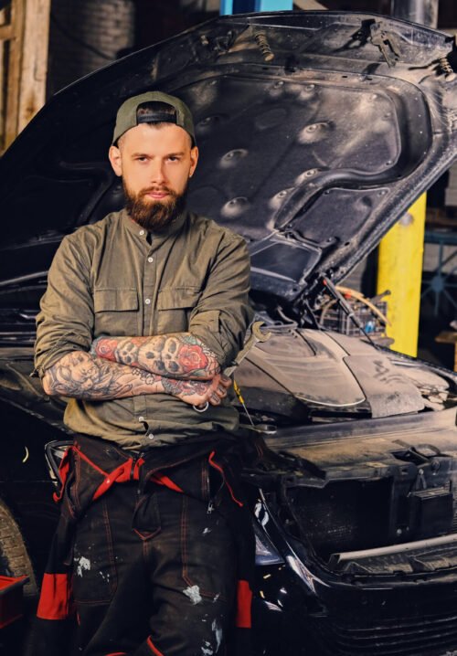 Portrait of bearded tattooed mechanics with crossed arms posing near a car in a workshop.