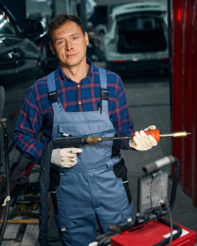 Portrait of professional man in protective costume standing near automobile, holding instrument and working at tire fitting
